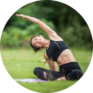 A smiling woman in black athletic wear performs a side stretch while seated on a yoga mat in a grassy outdoor setting.
